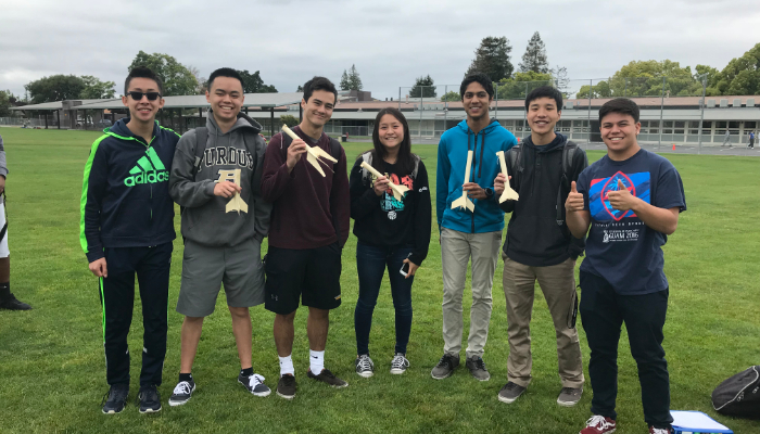 Physics students stand with handmade rocket parts in field readying for test launch